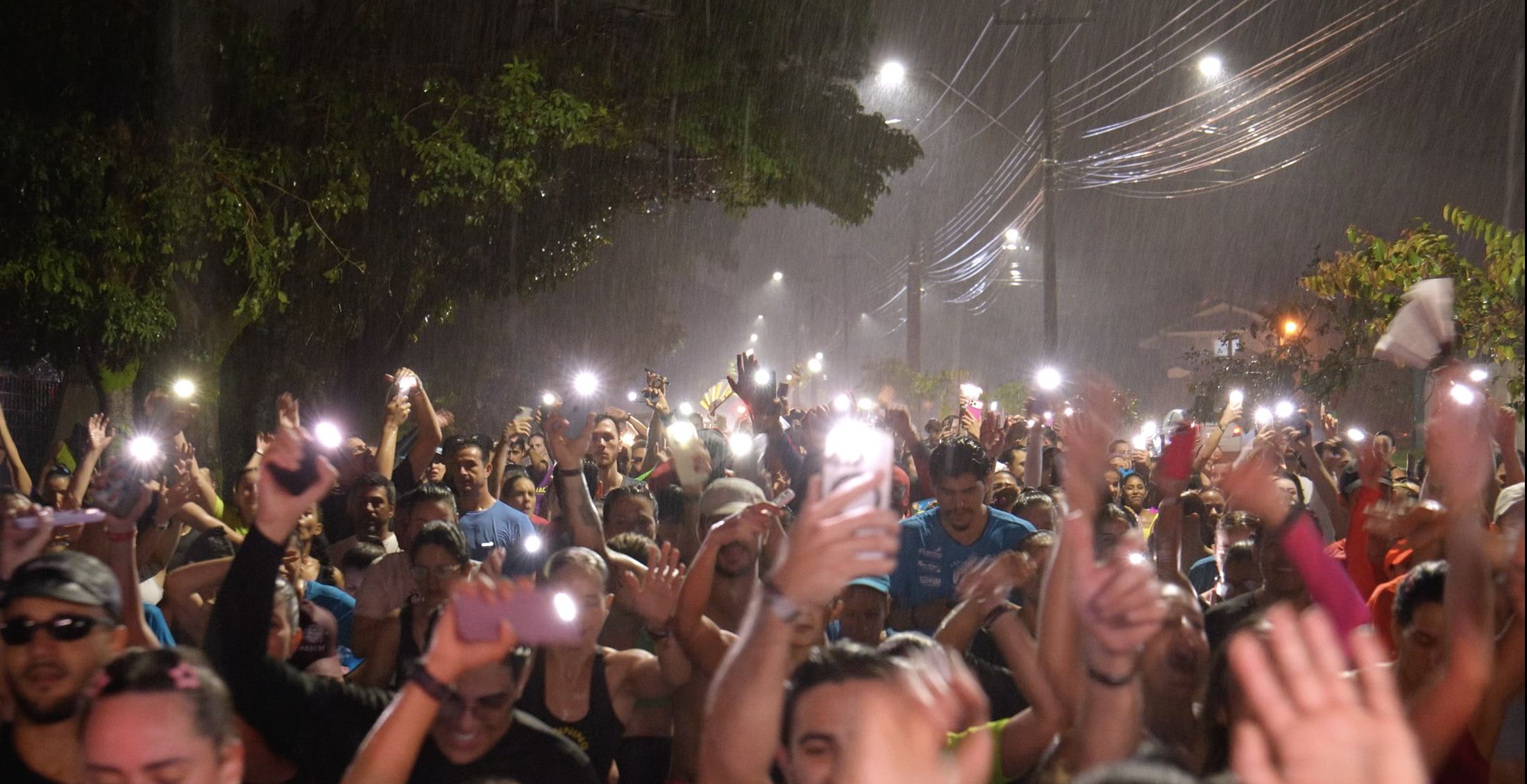 Multidão de corredores sob chuva à noite, com as lanternas dos celulares erguidas